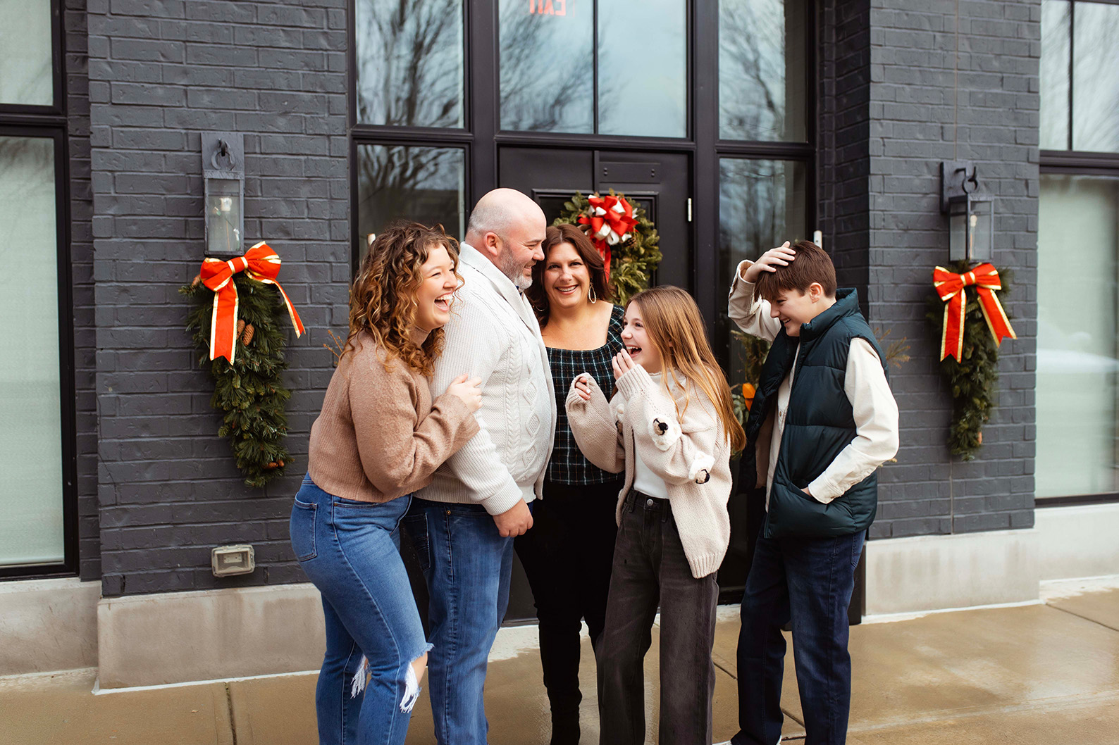  A family of 5 cuddled together in front of a building laughing. 