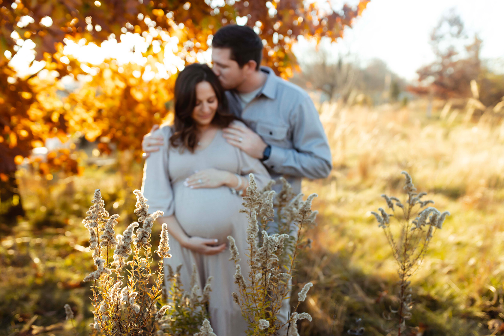 a pregnant woman holding her belly looking down, while her husband is behind her kissing her head