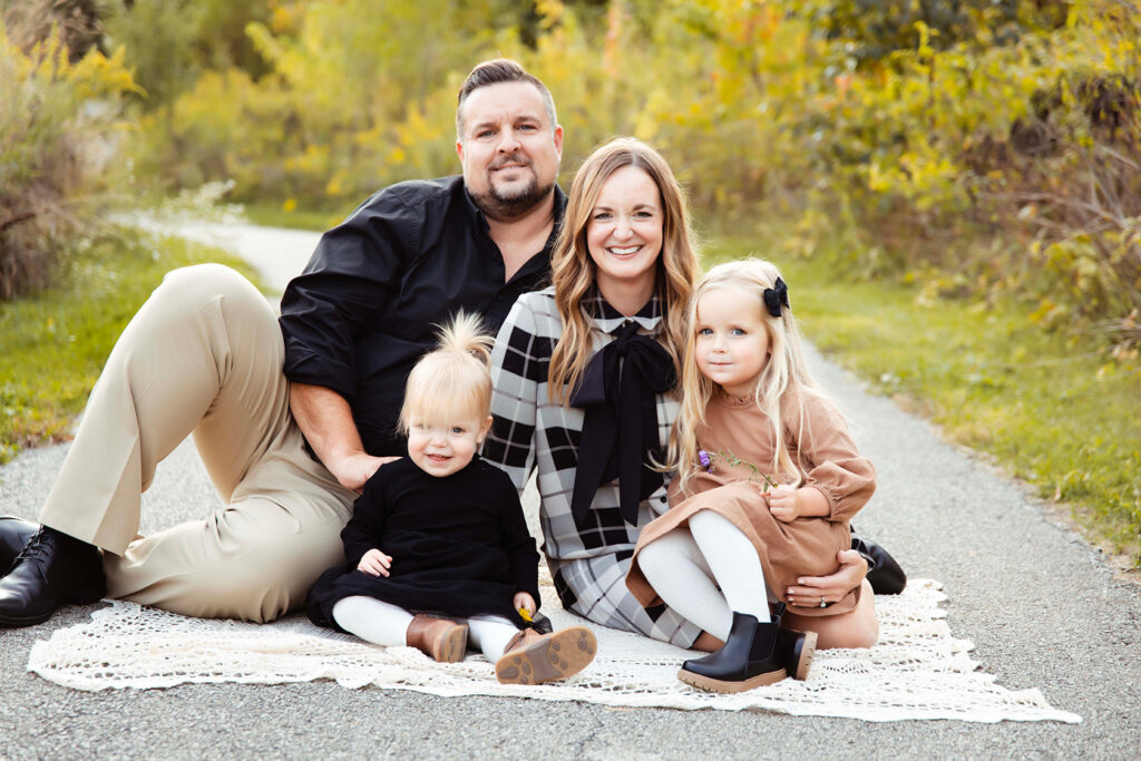 A family of 4 sitting on the ground together cuddling close and smiling. 