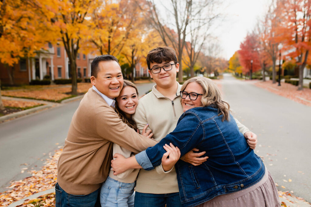 A family of four is hugging each other and smiling. The fall leaves are very colorful.