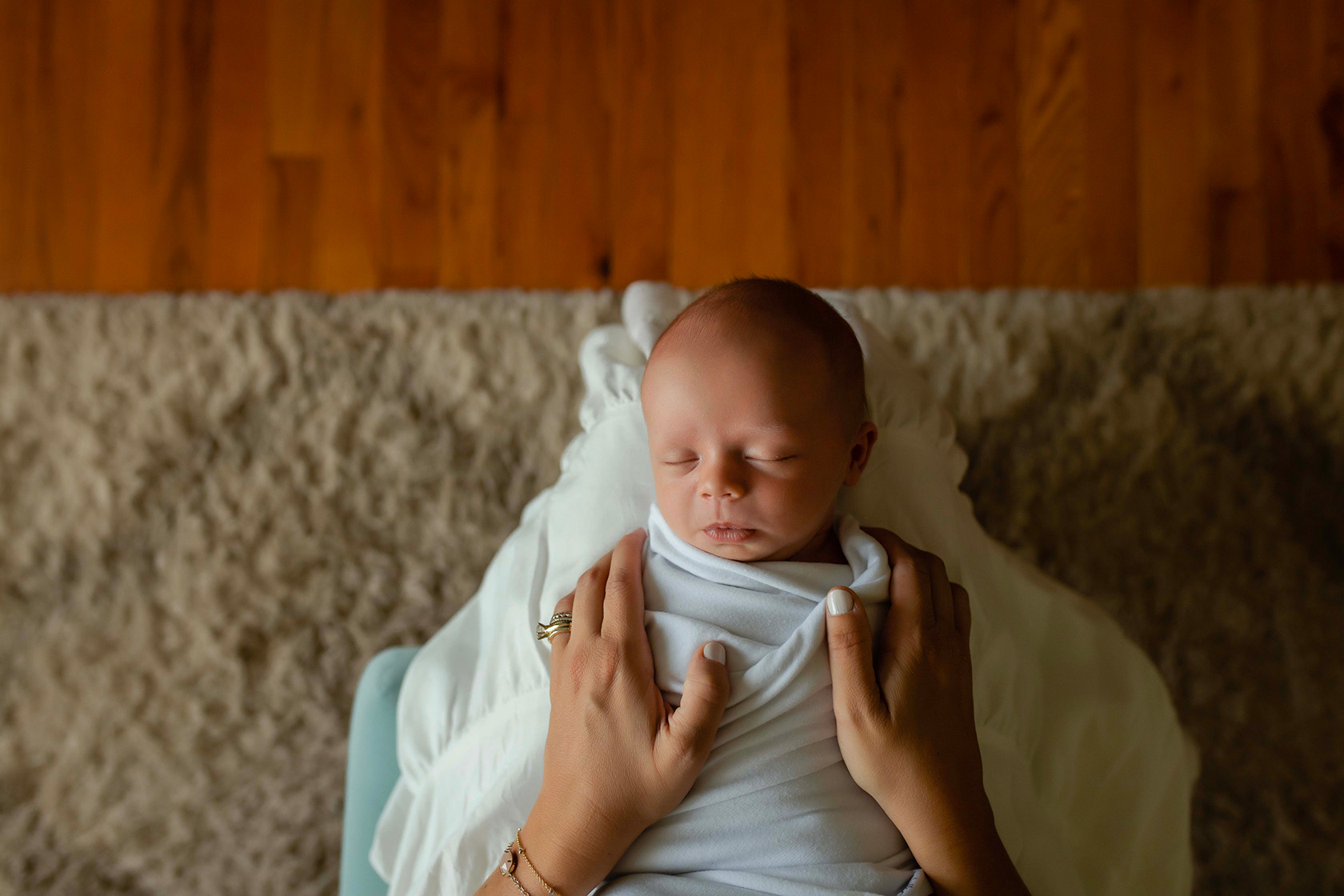 A baby at a newborn lifestyle session is wrapped in a white blanket laying on the moms lap. Mom has her hands around the baby. 