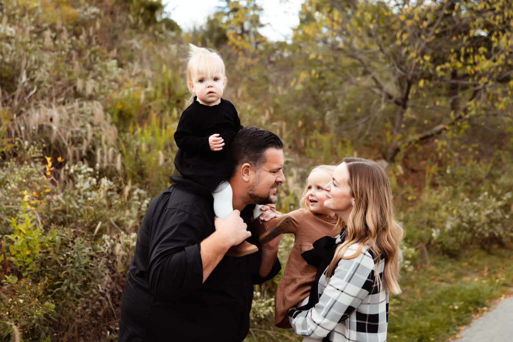 A little girl is up on her dad's shoulders, and her mom is holding her sister on her hip. Mom and Dad are going to kiss each other. 