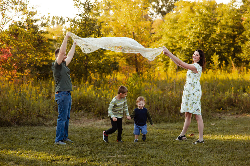 A mom and dad are lifting a blanket in the air while thier two boys run underneath holding hands. 