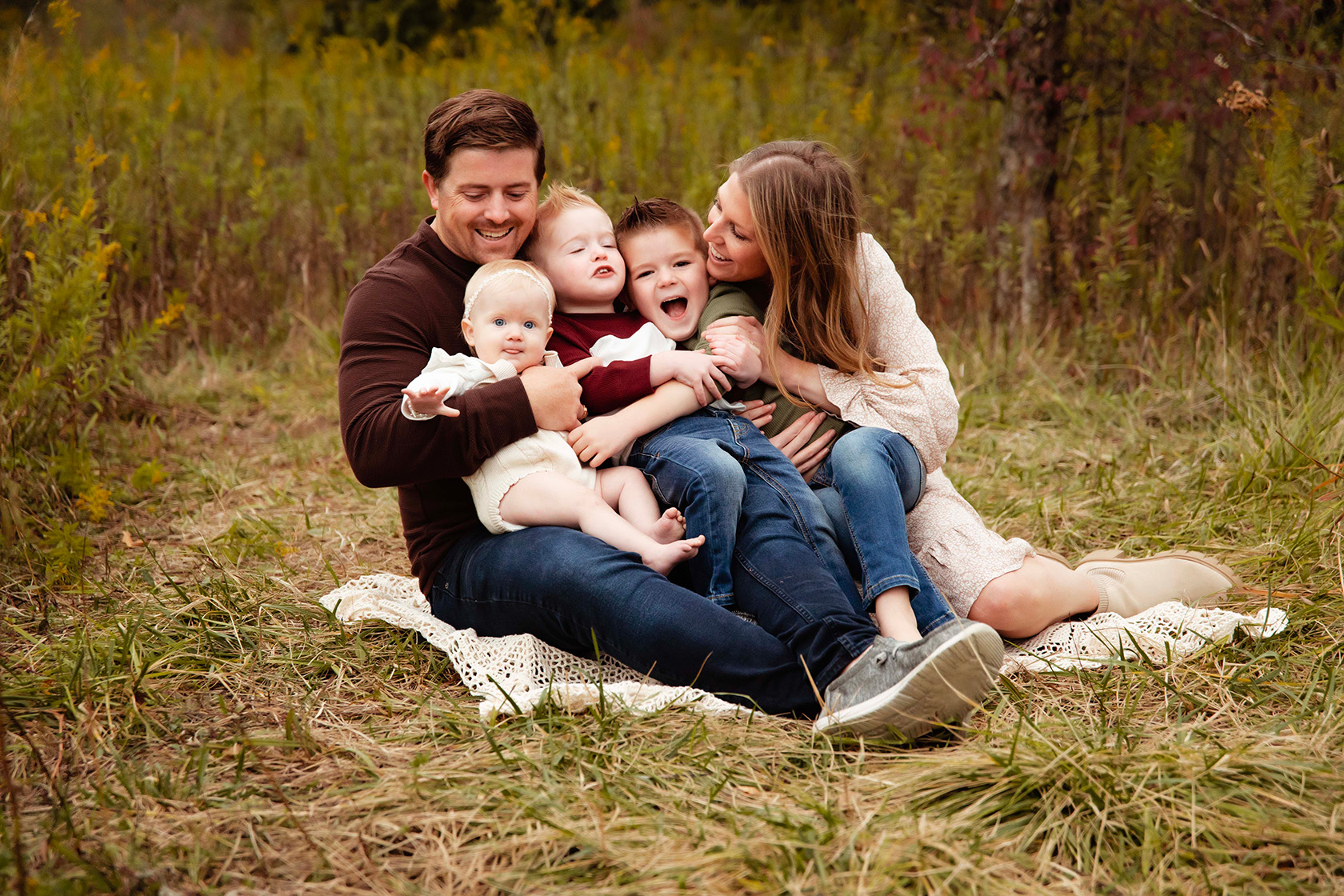 A family of 5 sits close together, having a tickle fight and laughing. 
