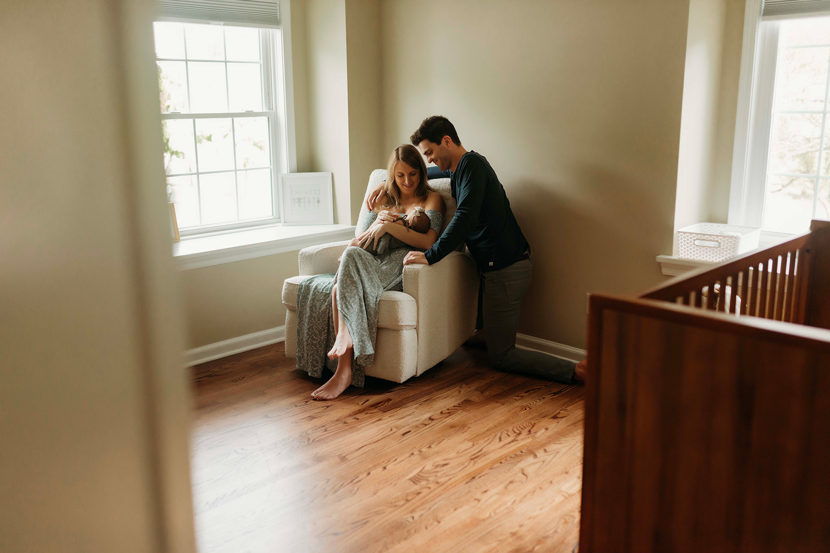 A mom is sitting in a rocking chair, holding her newborn baby, and her dad is kneeling down behind them, looking at the baby. 