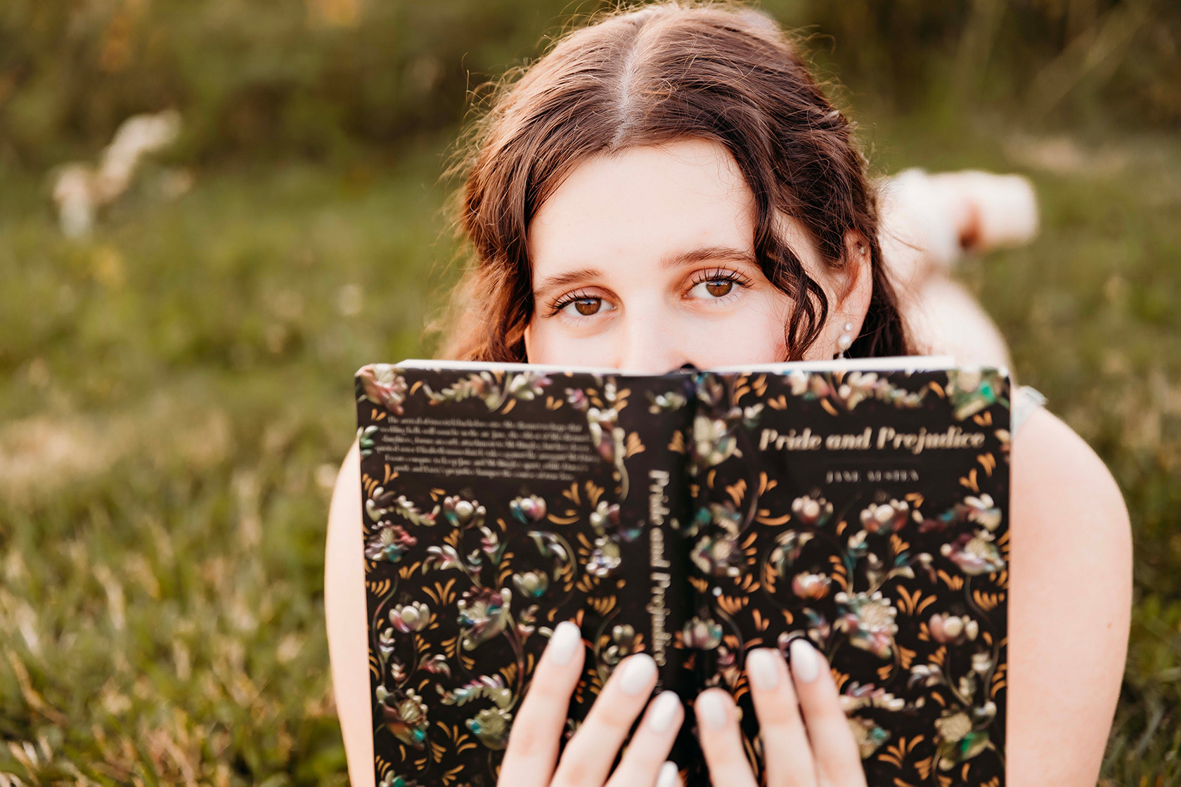 A girl laying on her belly, peaking over an open book for her senior portraits. 