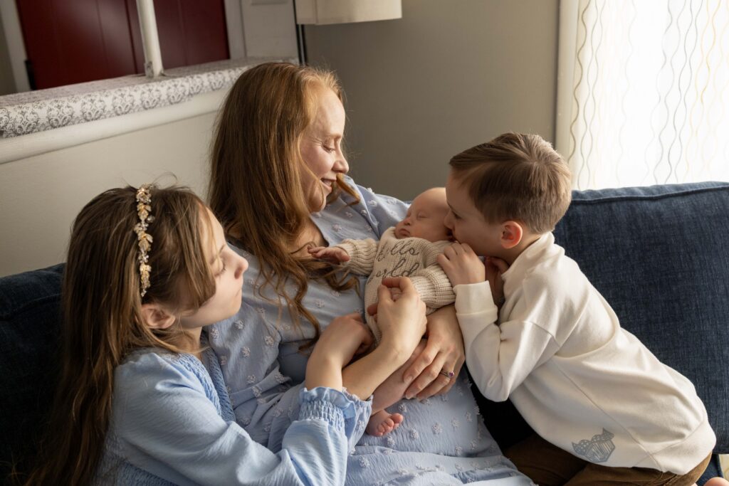 A mom is holding her newborn, sitting on a couch with her son and daughter. All looking at and loving on the baby. Carmel, Indiana Newborn photographer.