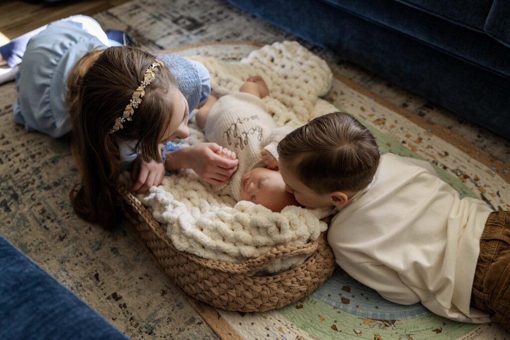 A newborn baby boy is on the floor in a little basket and chunky knit blanket. His sister and brother are lying close and giving him kisses.