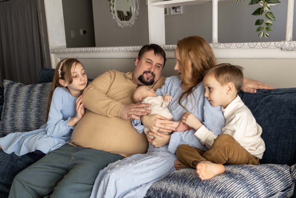A family of 5 is sitting on the couch snuggling the newest baby.