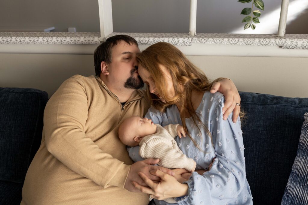 A mom and dad sitting together on the couch holding their newborn. Dad is kissing Mom on the head.