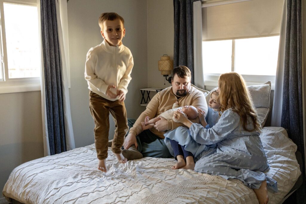 A family of 5 snuggled on a bed, snuggling a newborn baby while big brother jumps and smiles.