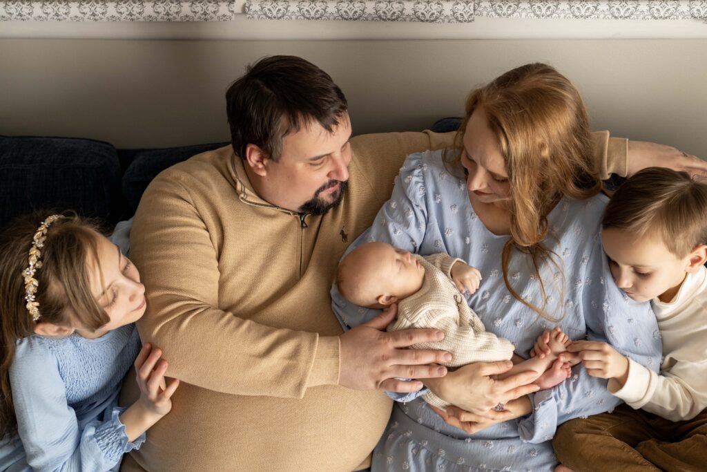 A mom is holding her new baby, sitting on a couch with her husband, son, and daughter. They're looking at the baby. Indianapolis newborn photographer.
