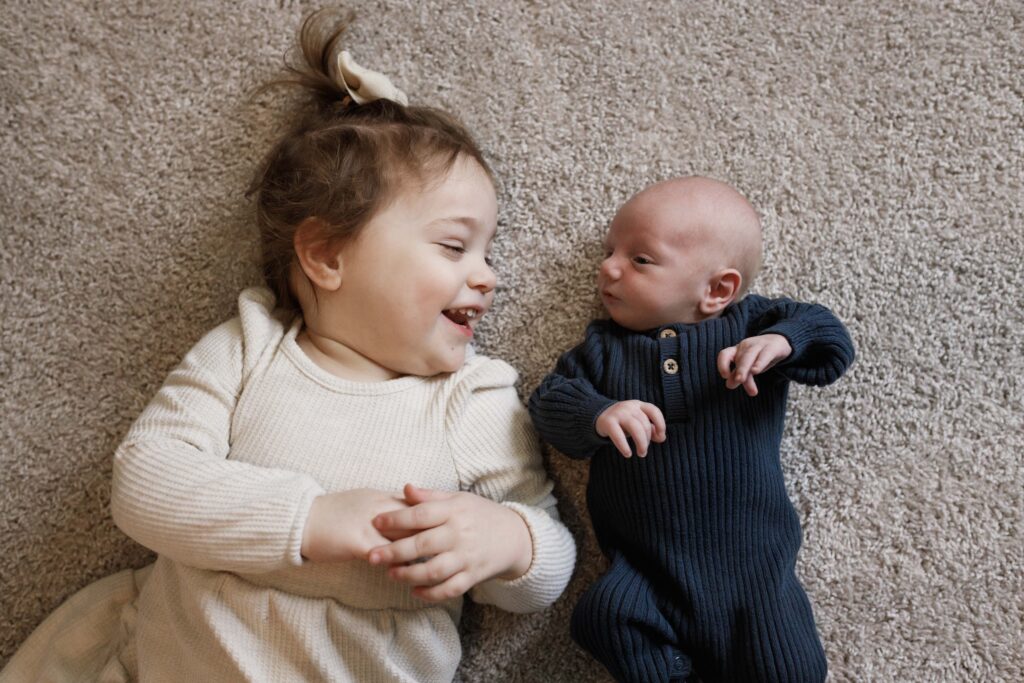 A little girl and her newborn brother lying on the floor looking at each other. 
