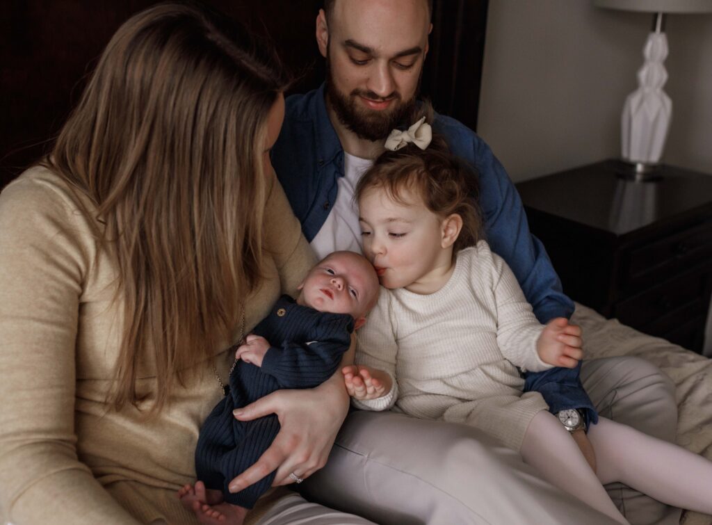 A mom is holding her newborn, and Dad is holding a toddler while she kisses the baby. They're all snuggled on a bed. Noblesville newborn photographer
